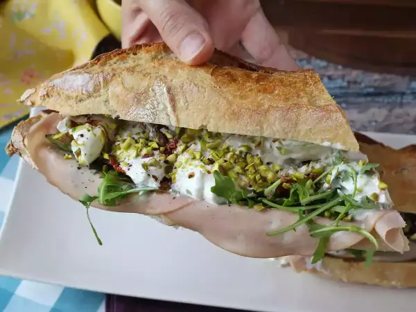 Bocadillo de pan rústico relleno de mortadela, burrata cremosa, rúcula, pistachos y tomates secos.