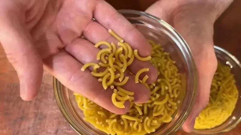 Mano sosteniendo fideos cortos y curvos sobre un bol de fideos similares.